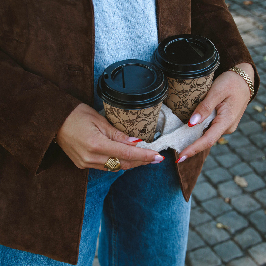 Hedda Huggie Ring i guld stylad på hand som håller två kaffekoppar. Statementring från Alora Avenue med elegant, vågformad design – vattentålig och tidlös.
