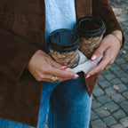 Hedda Huggie Ring i guld stylad på hand som håller två kaffekoppar. Statementring från Alora Avenue med elegant, vågformad design – vattentålig och tidlös.