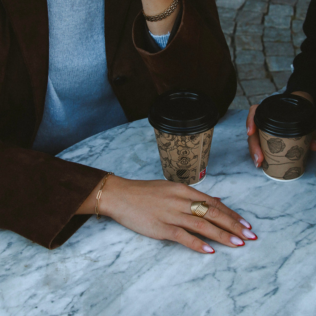 Hedda Huggie Ring i guld stylad på hand med kaffekopp på marmorbord. Elegant, vågformad ring från Alora Avenue – vattentålig, allergivänlig och tidlöst lyxig.