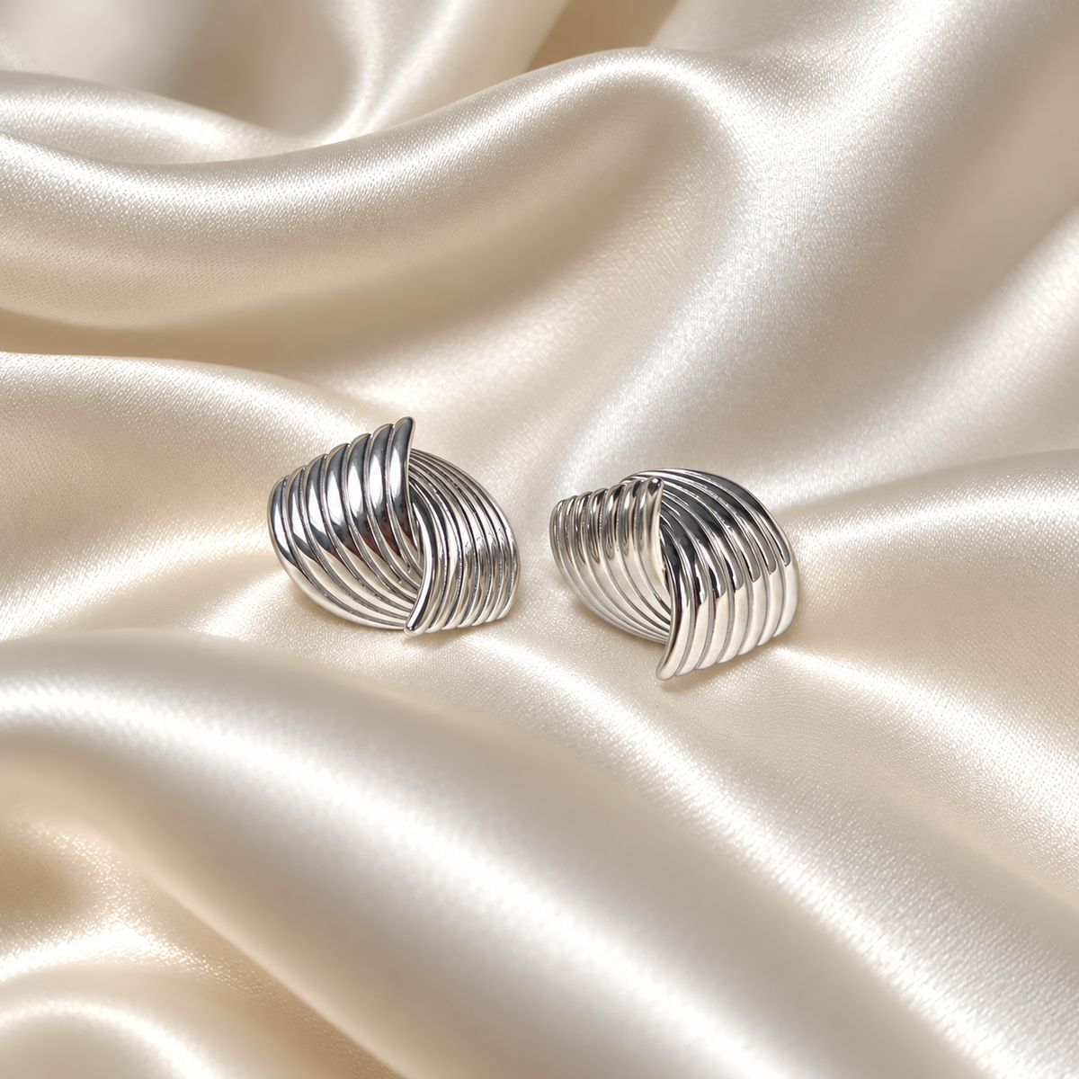 Holly Huggie Earrings i silver fotograferad på ljust silkestyg. Vattentåligt och allergivänligt smycke i rostfritt stål från Alora Avenue.