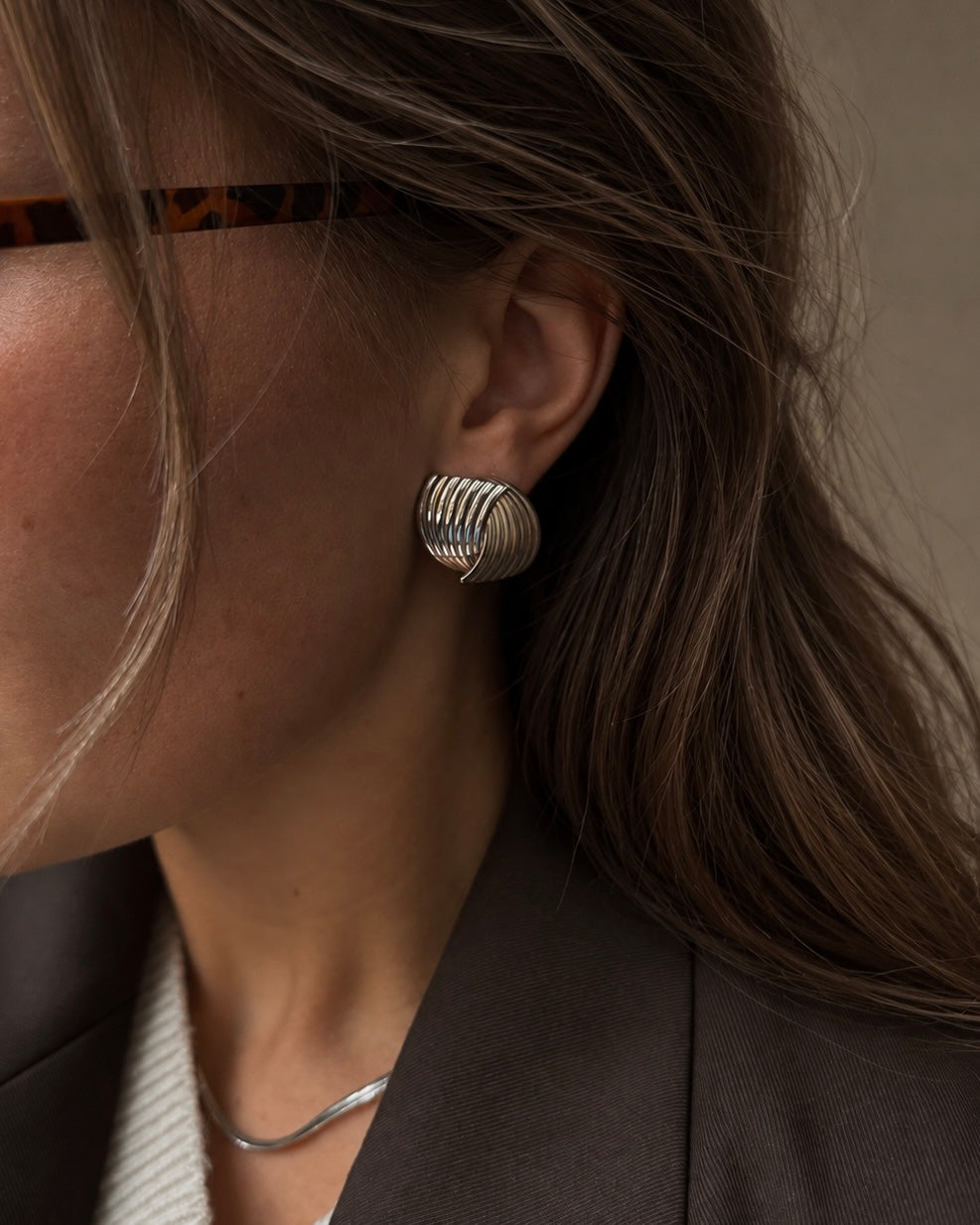 Holly Huggie Earrings i silver på modell – skulpterade statementörhängen med räfflat, vågigt mönster, burna med Cleo Cobra Necklace. Vattentåliga, allergivänliga örhängen i rostfritt stål från Alora Avenue.