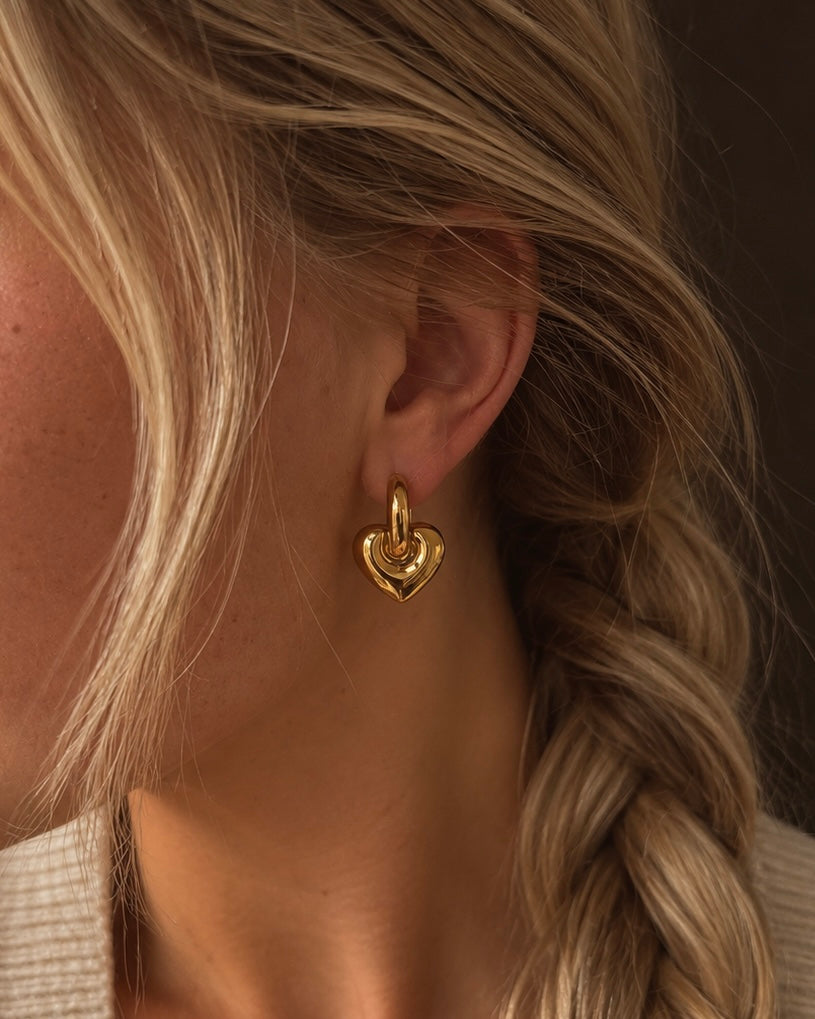 Lova Love Loops i guld på modell – hjärt-hoops med ett hängande hjärta i högblank finish. Vattentåliga, allergivänliga hjärtörhängen i rostfritt stål med 18K guldplätering från Alora Avenue.