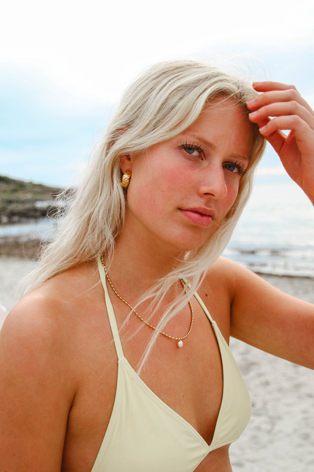Alora Avenue-smycken vid havet – Marigold Chunky Hoops och Polly Pearl Necklace i guld med pärla, bärs av modell på stranden. Vattentåliga och allergivänliga smycken för en avslappnad men lyxig sommarstil.