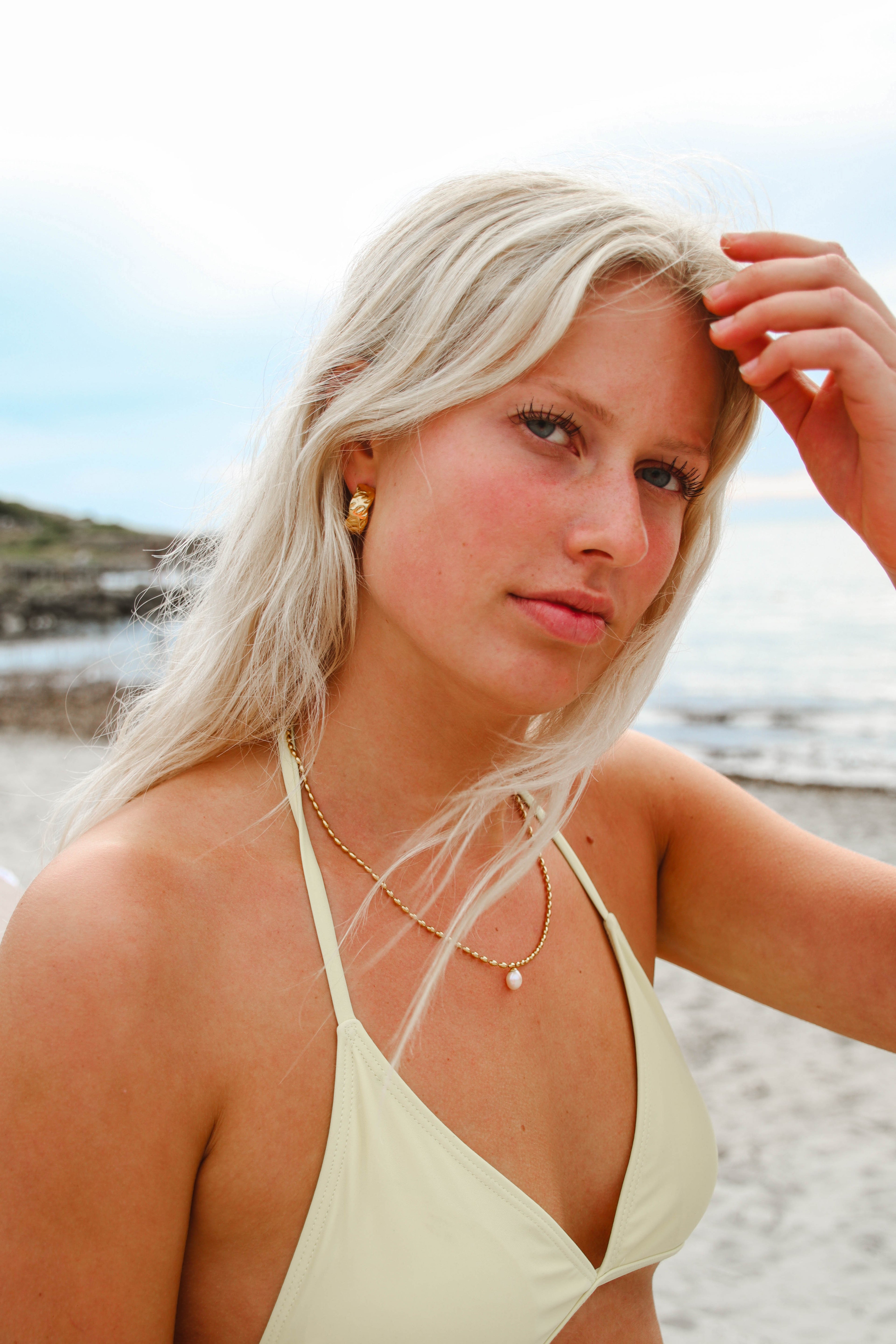 Alora Avenue-smycken vid havet – Marigold Chunky Hoops och Polly Pearl Necklace i guld med pärla, bärs av modell på stranden. Vattentåliga och allergivänliga smycken för en avslappnad men lyxig sommarstil.