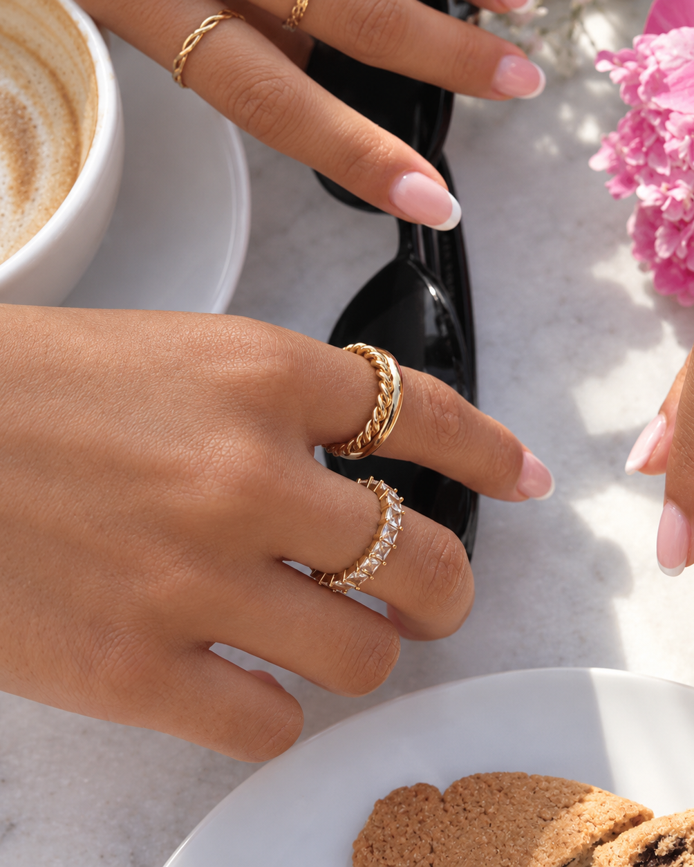 Minnie Moon Ring i guld buren i en ringstack på modell vid ett kaffebord i solljus. Vattentålig och allergivänlig ring i rostfritt stål med 18K PVD-guldplätering från Alora Avenue.