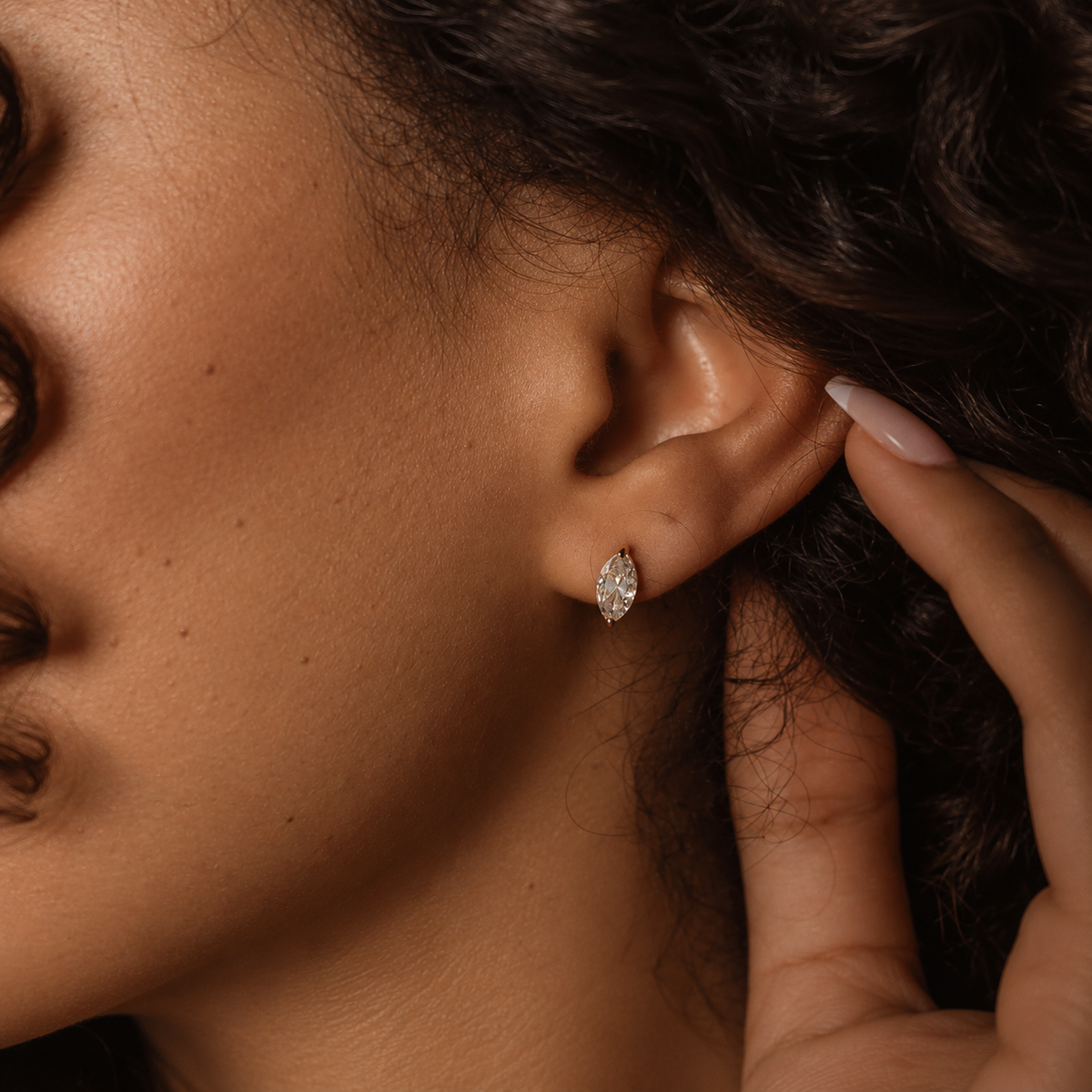 Nara Navette Earrings i guld på modell – närbild på örat med en navetteslipad vit sten i guldinfattning. Vattentåliga och allergivänliga örhängen i rostfritt stål med 18K PVD-guldplätering från Alora Avenue.