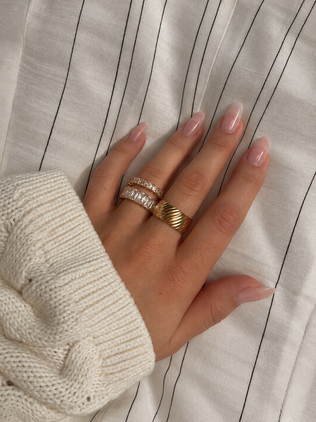 Oblique Orion Ring i guld buren i en ringstack på modell mot en mjuk stickad tröja. Vattentålig och allergivänlig ring i rostfritt stål med 18K PVD-guldplätering från Alora Avenue.