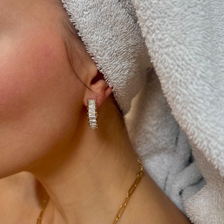 Närbild på Sabrina Spark Hoops från Alora Avenue – eleganta guldörhängen med gnistrande zirkoner, stylade med en handduk runt håret och matchande halsband i guld.