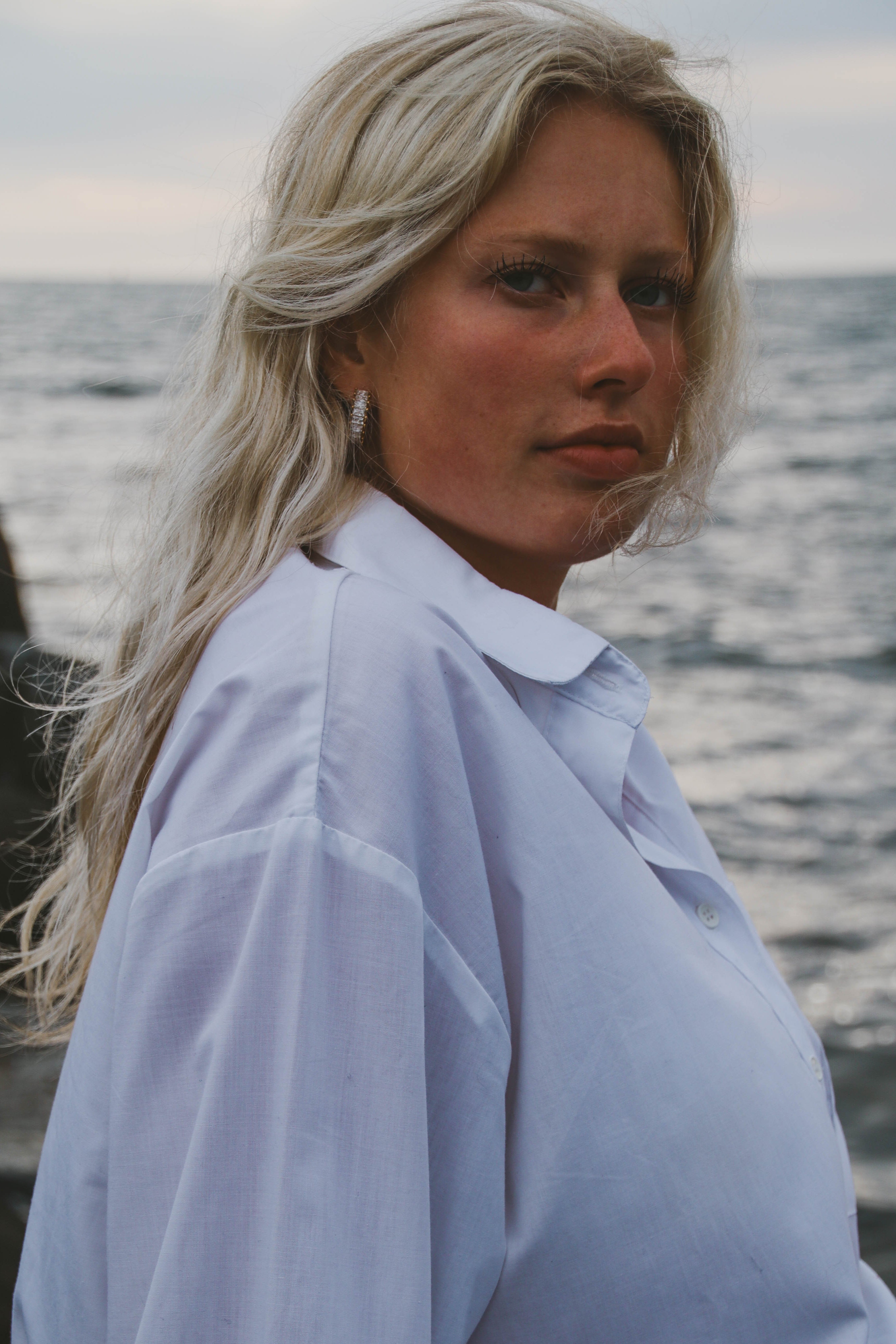 Närbild på modell i vit skjorta vid havet som bär Alora Avenue's glittrande Sabrina Spark Hoops – eleganta guldörhängen perfekta för både vardag och fest. Vattentåliga, allergivänliga smycken med tidlös lyster.