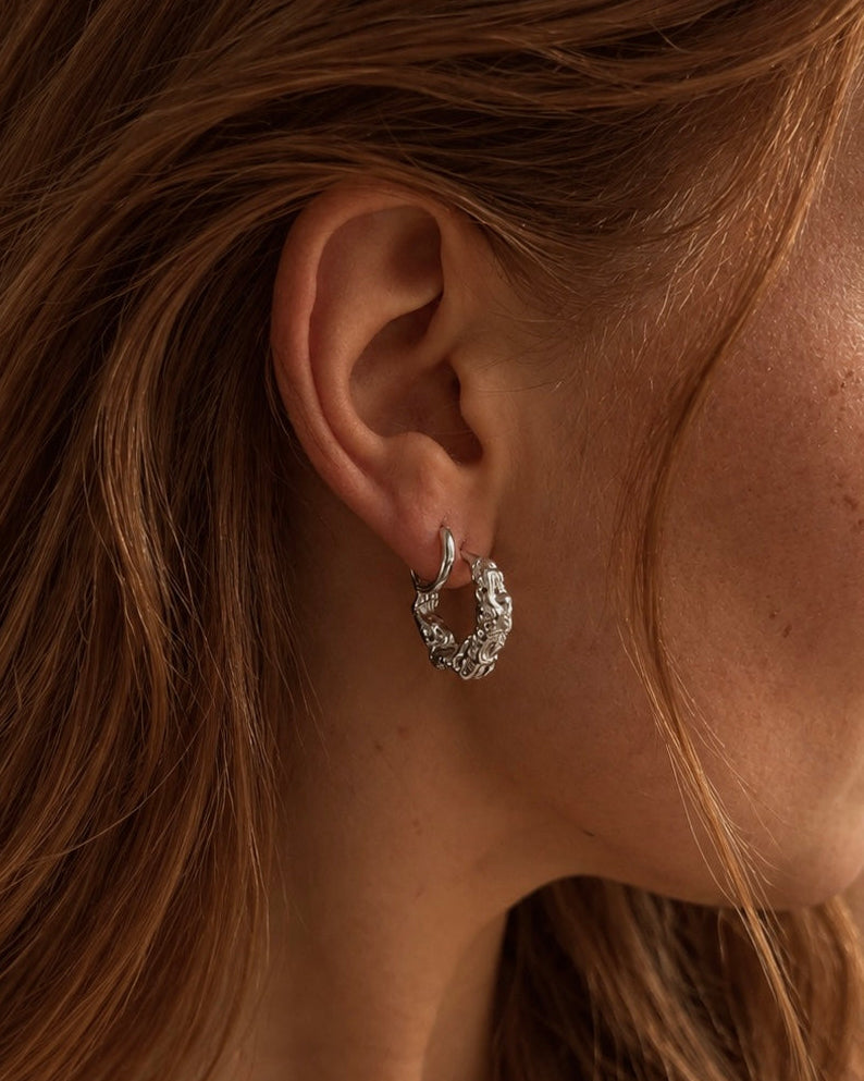 Saga Sway Hoops i silver på modell – chunky hoops med organisk, hamrad struktur som fångar ljuset. Vattentåliga, allergivänliga örhängen i rostfritt stål från Alora Avenue.