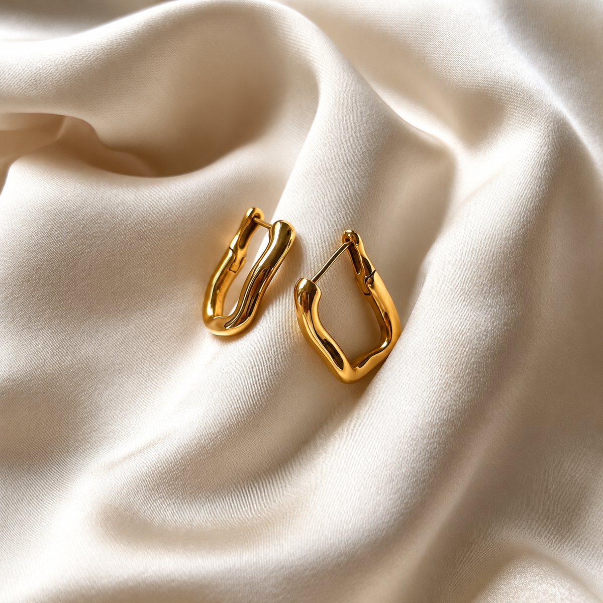 Saint Poppy Hoops i guld fotograferade på ljust silkestyg. Vattentåliga och allergivänliga örhängen i rostfritt stål med 18K PVD-guldplätering från Alora Avenue.