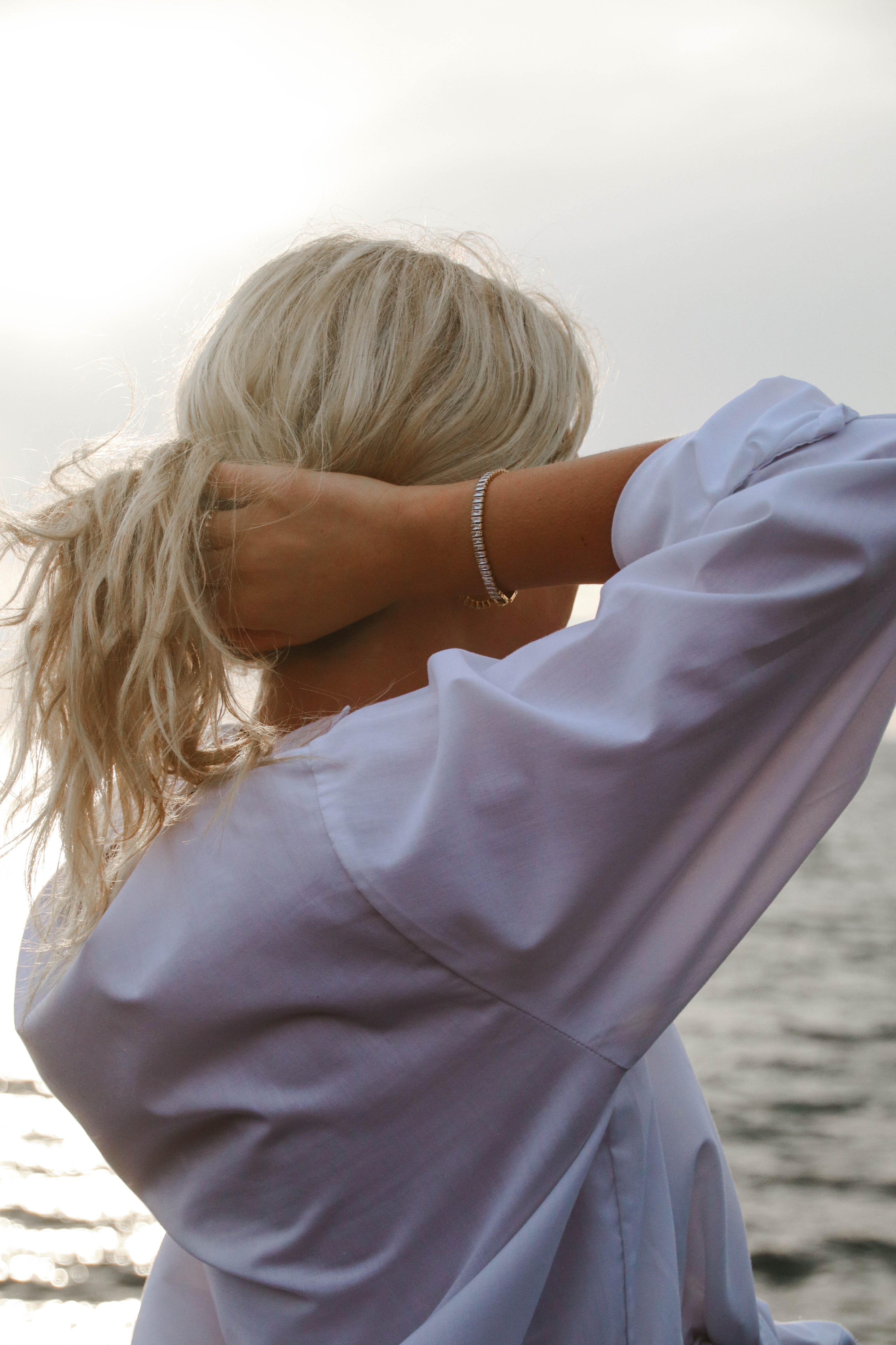 Santiago Sunstone Bracelet från Alora Avenue stylat vid havet – ett glittrande armband i guld med baguetteslipade stenar, buren med vit skjorta i solnedgången.