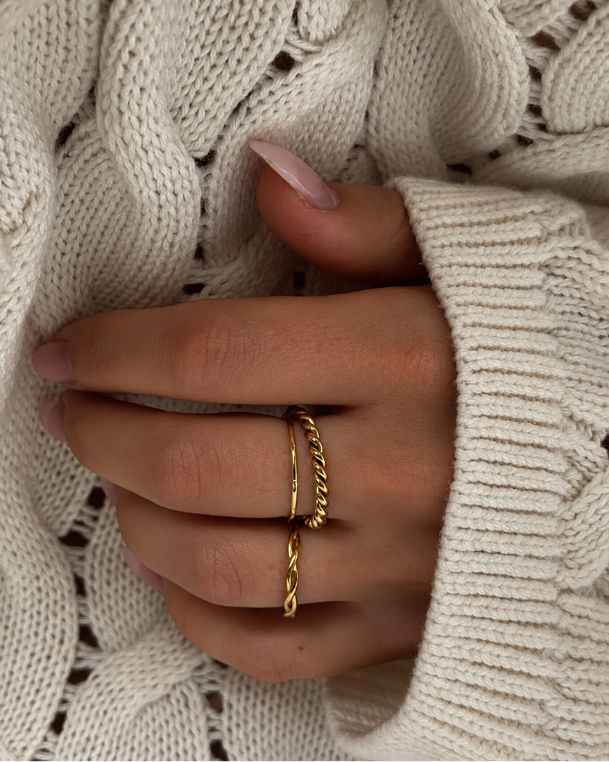 Serpentine Sea Ring i guld på modell mot en kabelstickad tröja, buren tillsammans med en kedjering. Vattentålig och allergivänlig ring i rostfritt stål med 18K PVD-guldplätering från Alora Avenue.