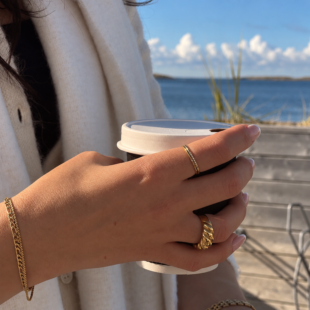 Shim Shim Ring i guld på modell som håller en kaffemugg vid havet. Vattentålig och allergivänlig ring i rostfritt stål med 18K PVD-guldplätering från Alora Avenue.
