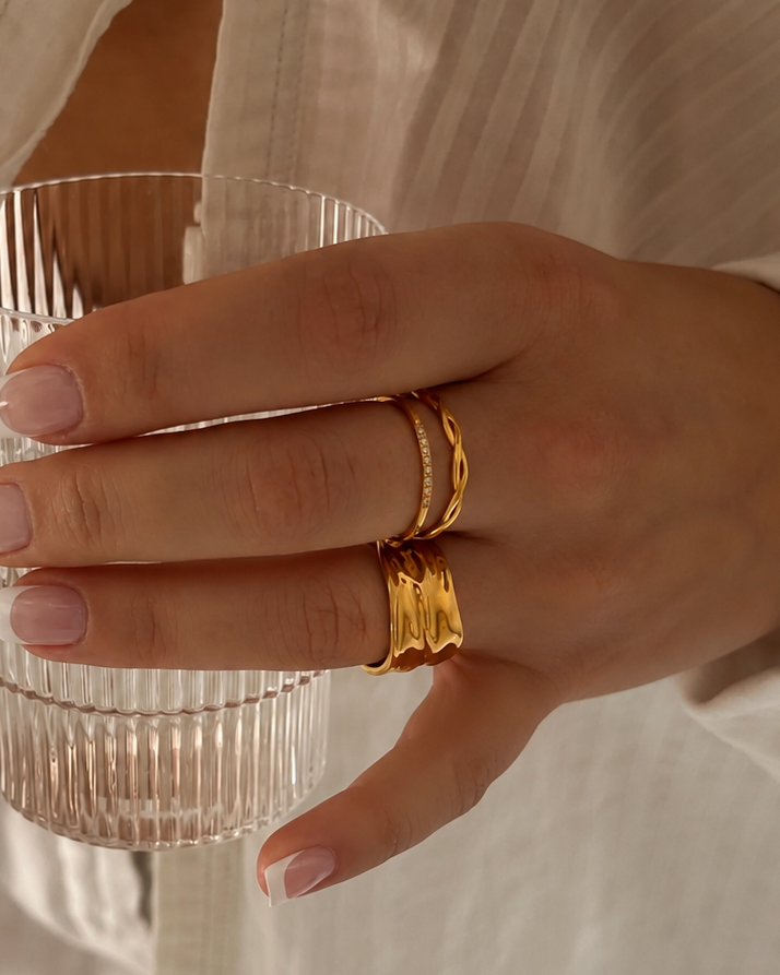 Shim Shim Ring i guld buren i en ringstack på modell som håller ett räfflat glas. Vattentålig och allergivänlig ring i rostfritt stål med 18K PVD-guldplätering från Alora Avenue.