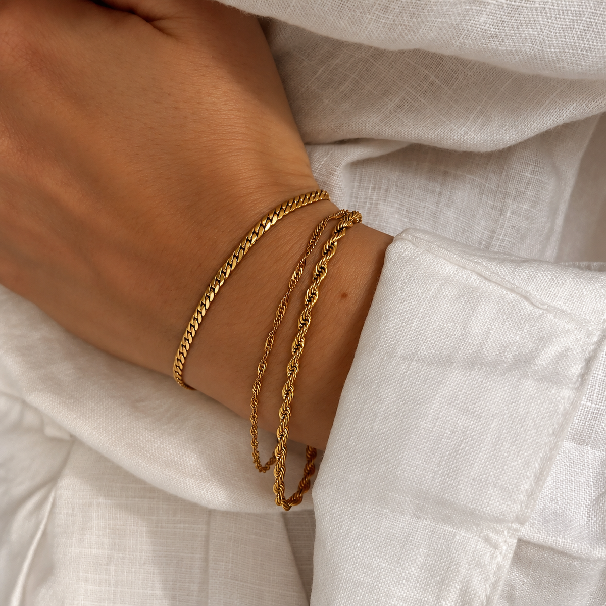 Sia Singapore Bracelet i guld buret i en armbandsstack på modell mot vitt linne. Vattentåligt och allergivänligt armband i rostfritt stål med 18K PVD-guldplätering från Alora Avenue.