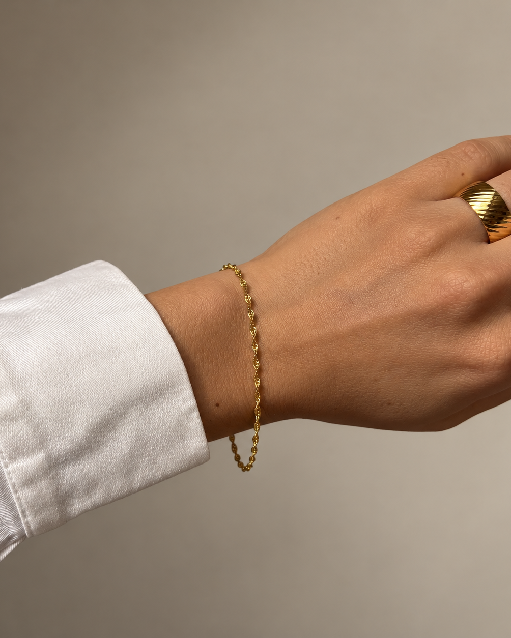 Sia Singapore Bracelet i guld på modellens handled till vit skjorta – tunn tvinnad kedja, stylad med en guldring. Vattentåligt och allergivänligt armband i rostfritt stål med 18K PVD-guldplätering från Alora Avenue.