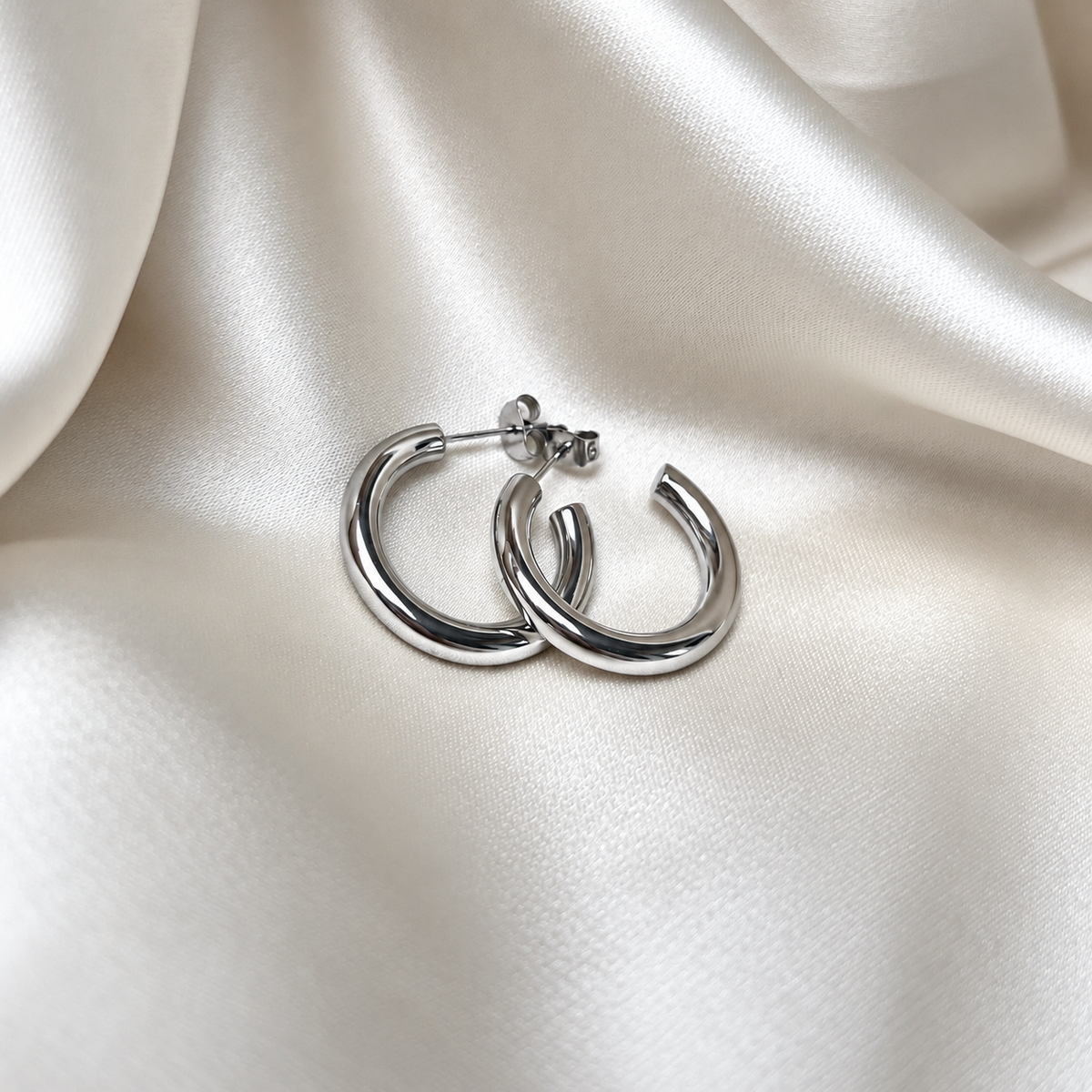 Simple Dakota Hoops i silver fotograferade på ljust silkestyg. Vattentåliga och allergivänliga örhängen i rostfritt stål från Alora Avenue.