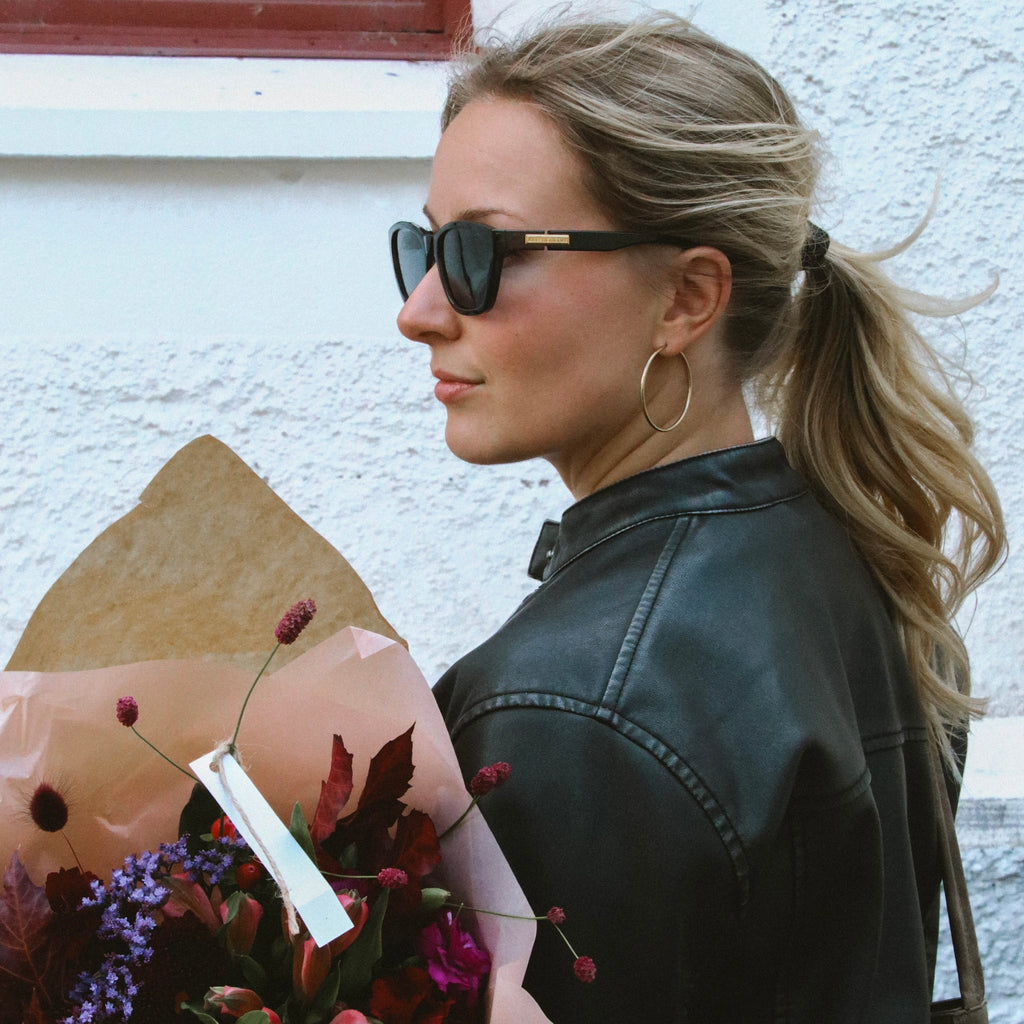 Sky Saturn Hoops i guld burna av kvinna i skinnjacka med blombukett. Stora, eleganta hoops från Alora Avenue – vattentåliga och perfekta för vardag och fest.