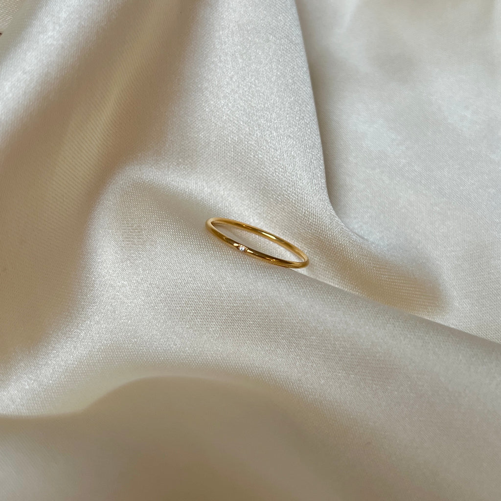 Solo Dazzle Ring på champagnefärgad siden – minimalistisk tunn guldfärgad ring med en liten klar sten. Vattentålig och allergivänlig, tillverkad i rostfritt stål med hållbar PVD-plätering. Perfekt för diskret vardagslyx eller att kombinera i en ringstack.