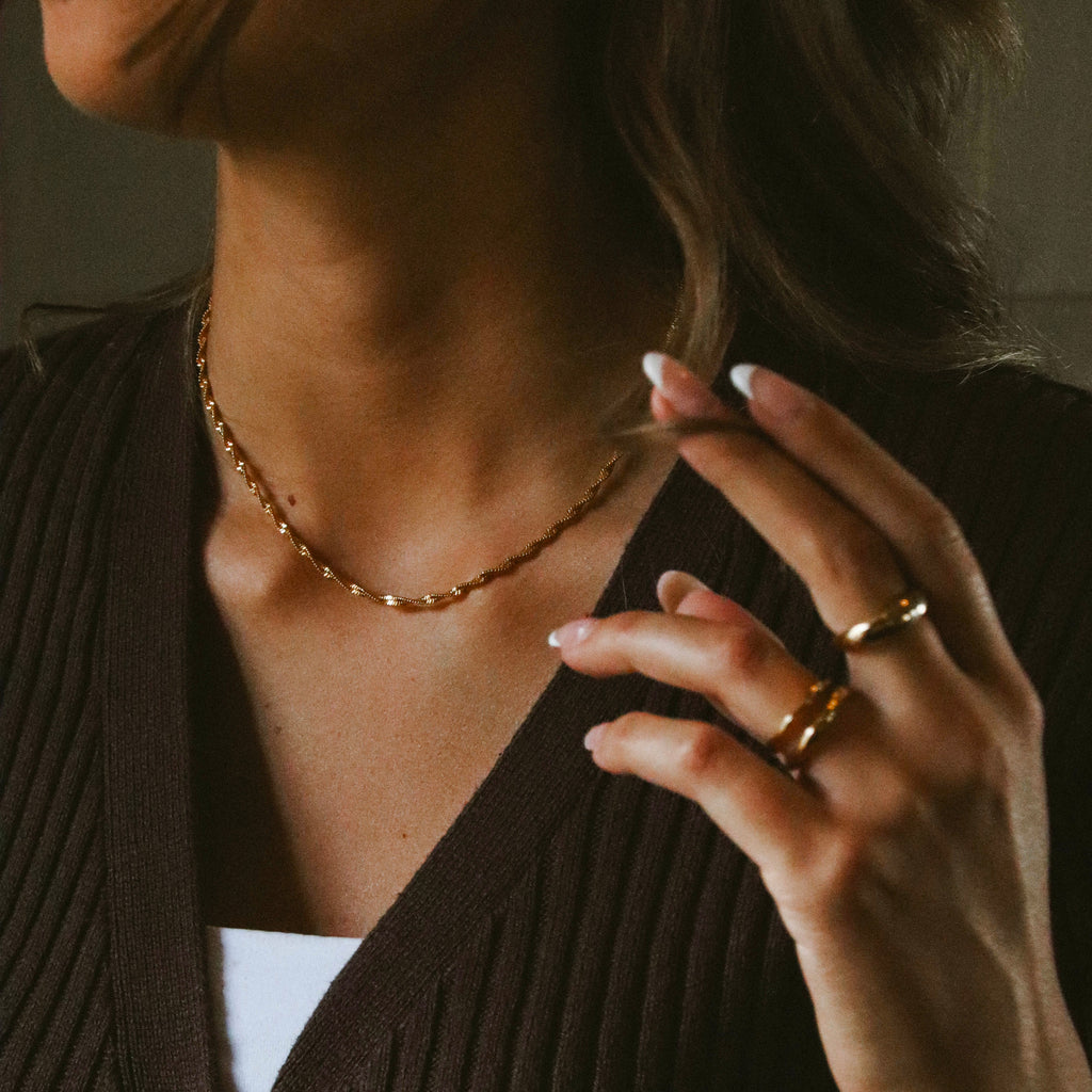 Tiana Twisted Necklace i guld på kvinna i brun tröja. Elegant tvinnat halsband från Alora Avenue – vattentåligt och perfekt för både vardag och fest.