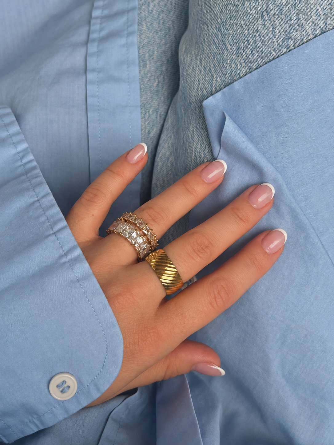 Tidy Crystal Ring i guld på modell mot en ljusblå skjorta, buren i en ringstack. Vattentålig och allergivänlig ring i rostfritt stål med 18K PVD-guldplätering från Alora Avenue.