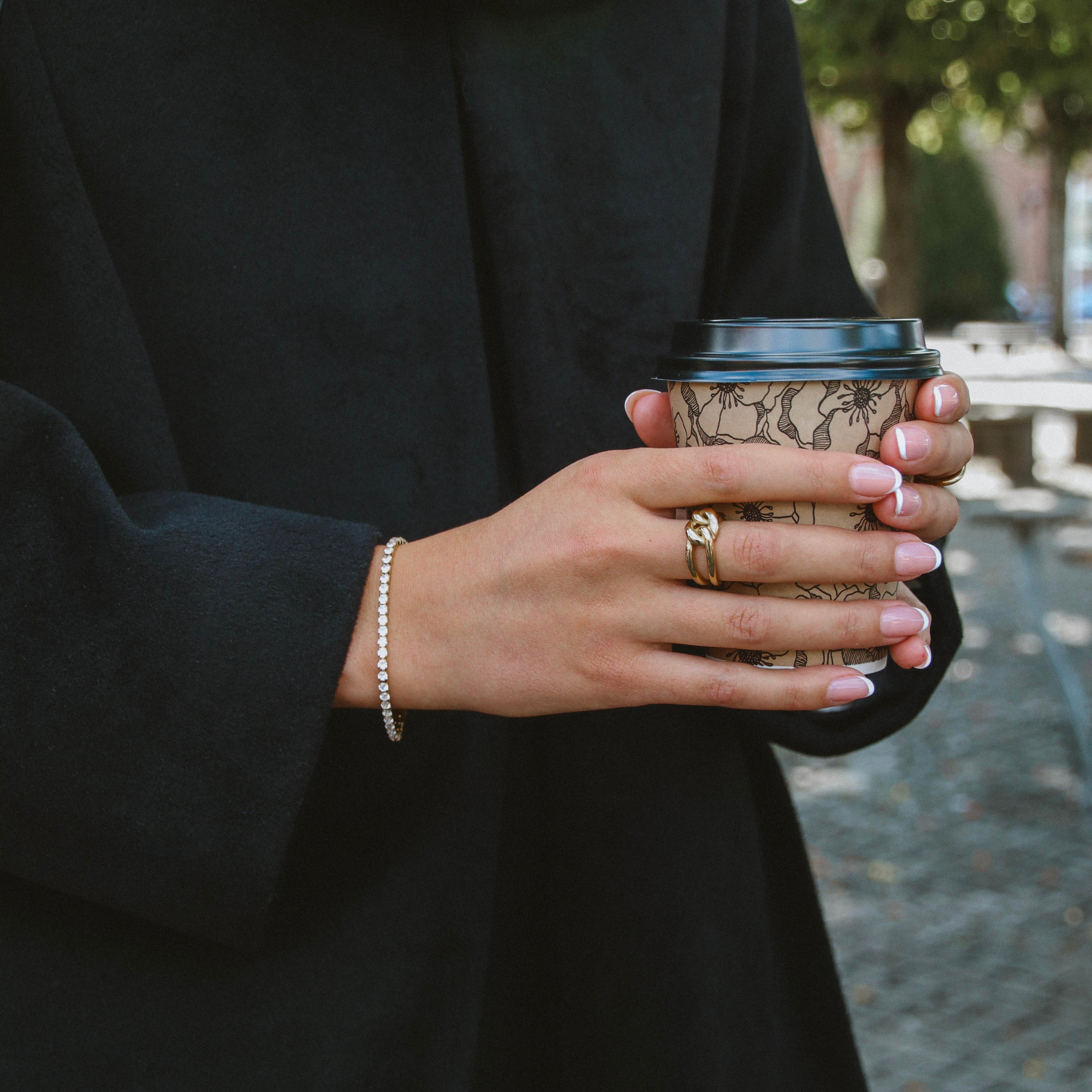 Tilda Tennis Bracelet i guld på handled, stylat med kaffekopp och svart kappa. Tidlöst armband med gnistrande stenar från Alora Avenue – vattentåligt och elegant.