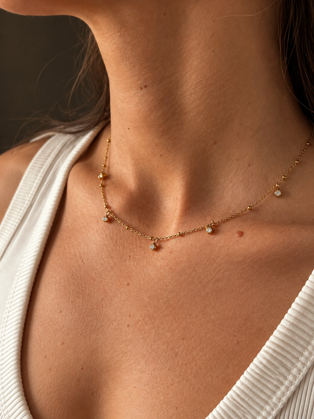 Vera V Necklace i guld med fem små vita stenar på en tunn kedja, buret på modell i vit ribbad topp. Vattentåligt och allergivänligt halsband från Alora Avenue.