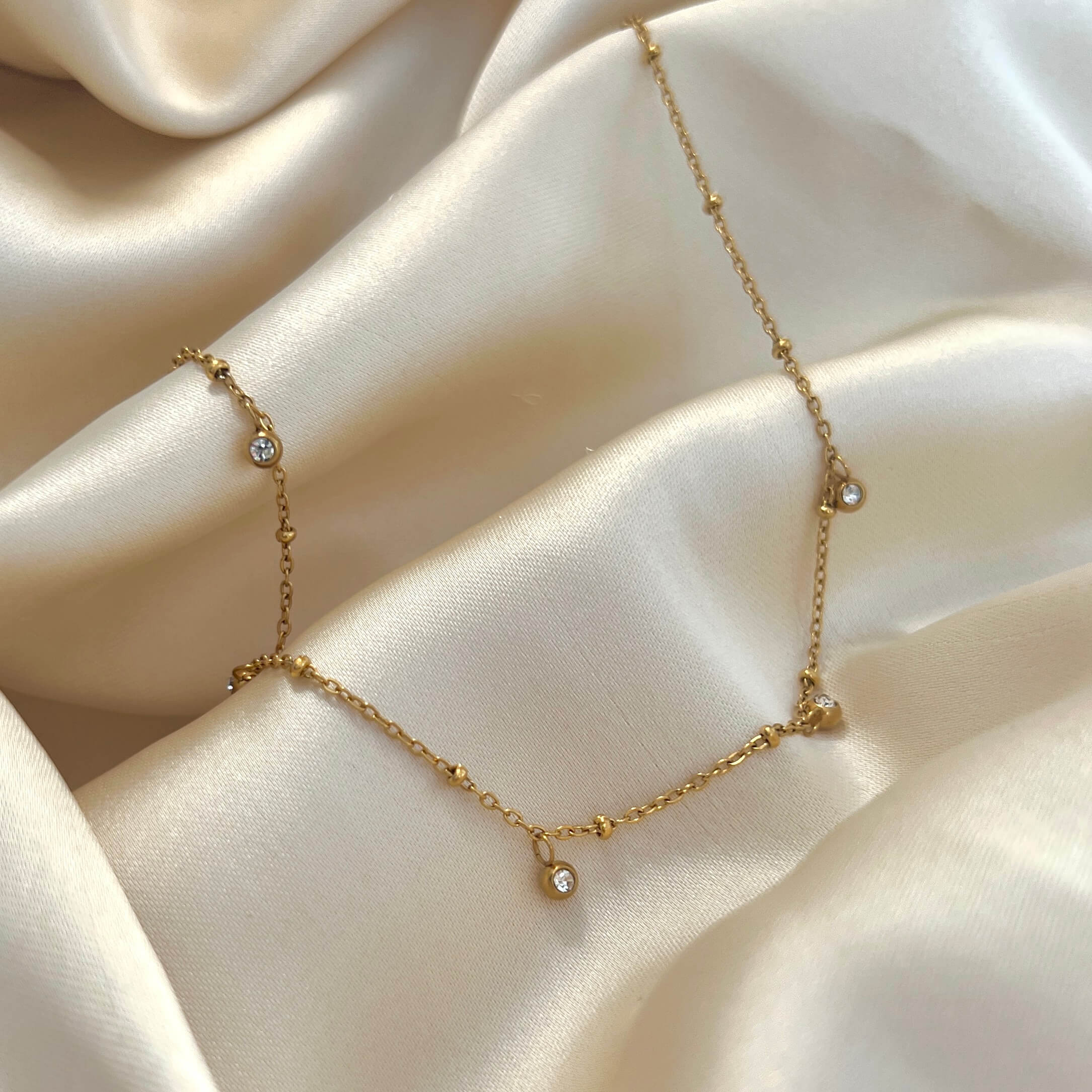 Vera V Necklace i guld med fem vita stenar fotograferat på ljus sidenbakgrund. Elegant och vattentåligt halsband från Alora Avenue – perfekt för layering eller solo.