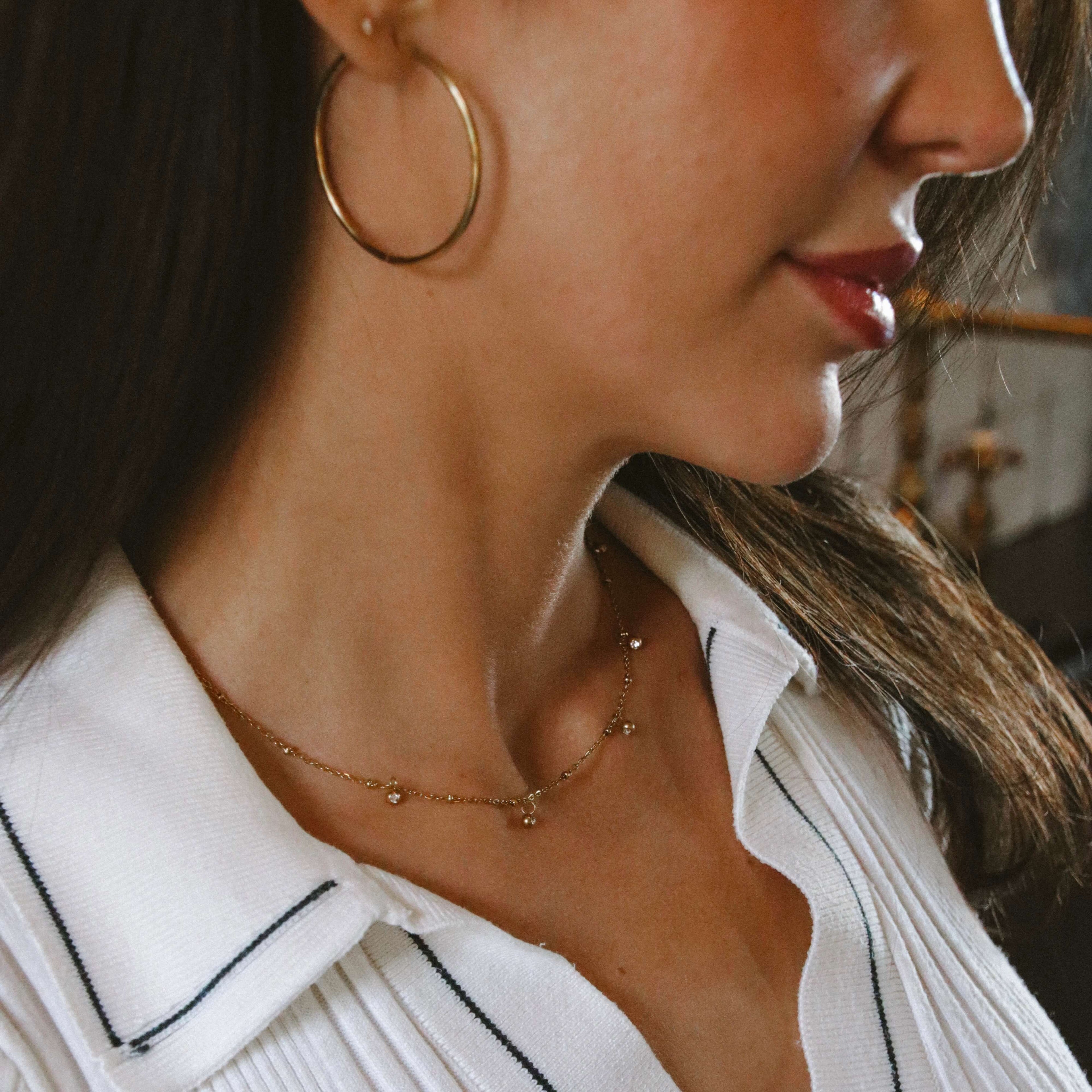 Guldhalsbandet Vera V Necklace med fem vita stenar stylat på modell i vit topp. Vattentåligt och färgbeständigt smycke från Alora Avenue – perfekt vardagslyx.