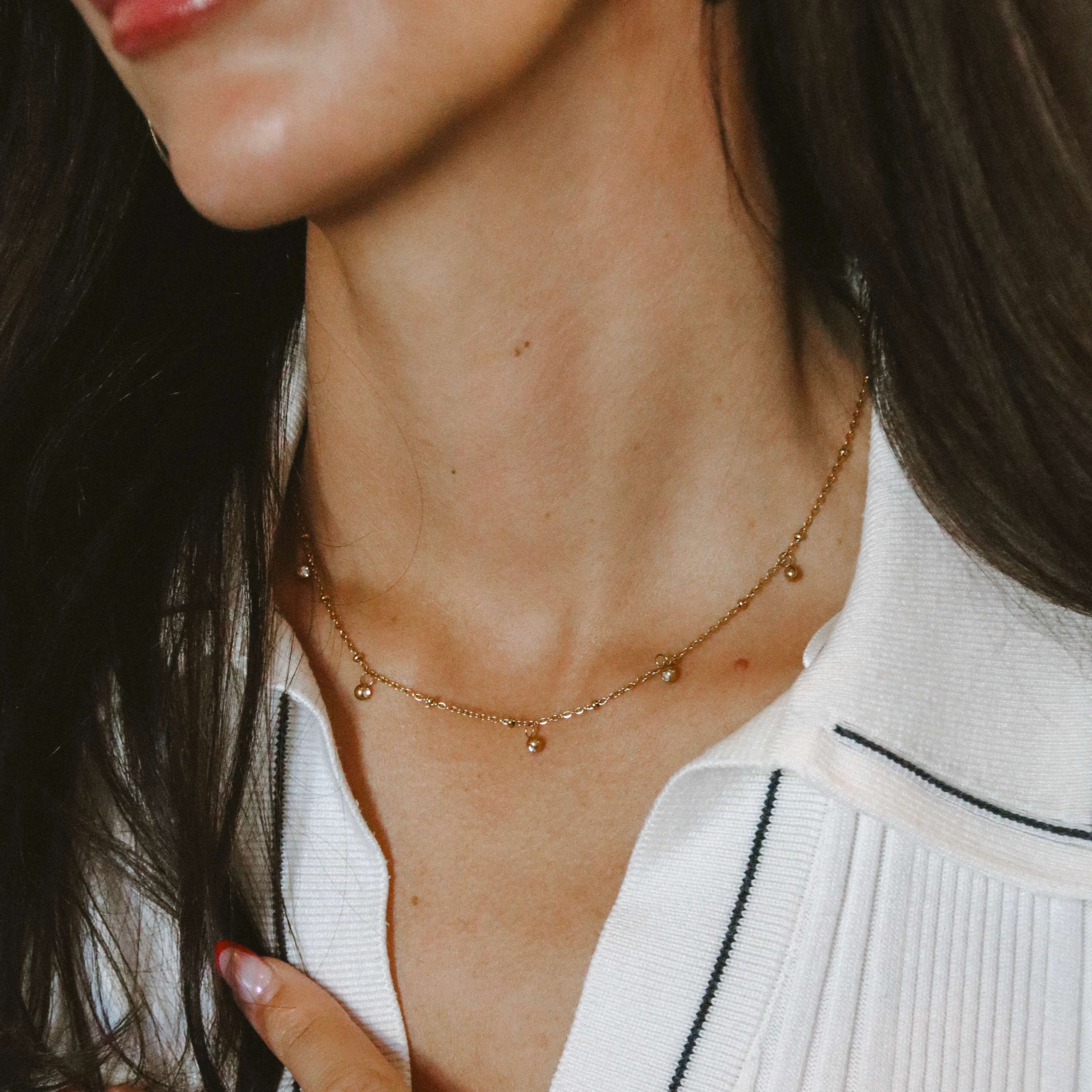 Vera V Necklace i guld med fem vita stenar, stylat på modell i vit topp. Tunt och elegant halsband från Alora Avenue – vattentåligt, allergivänligt och perfekt att bära varje dag.