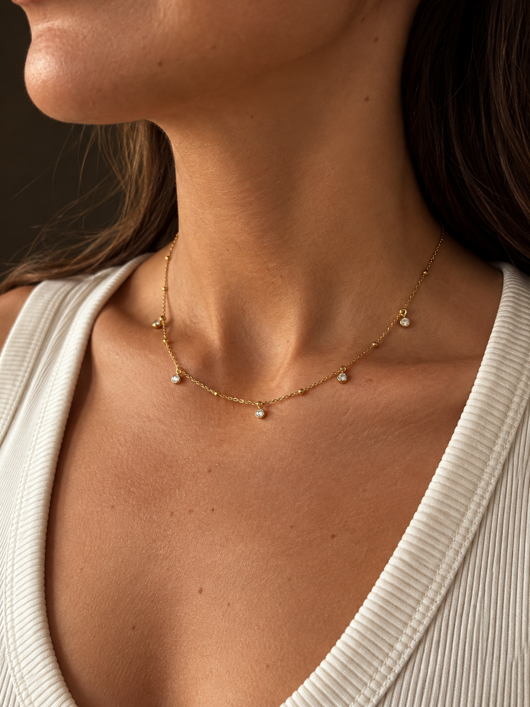 Närbild på Vera V Necklace i guld mot halsgropen, stylat till en vit ribbad v-ringad topp. Tunn kedja med fem glittrande stenar – vattentåligt och färgbeständigt smycke från Alora Avenue.