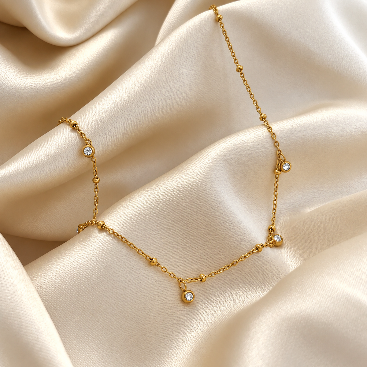 Vera V Necklace i guld fotograferad på ljust silkestyg. Vattentåligt och allergivänligt smycke i rostfritt stål med 18K PVD-guldplätering från Alora Avenue.