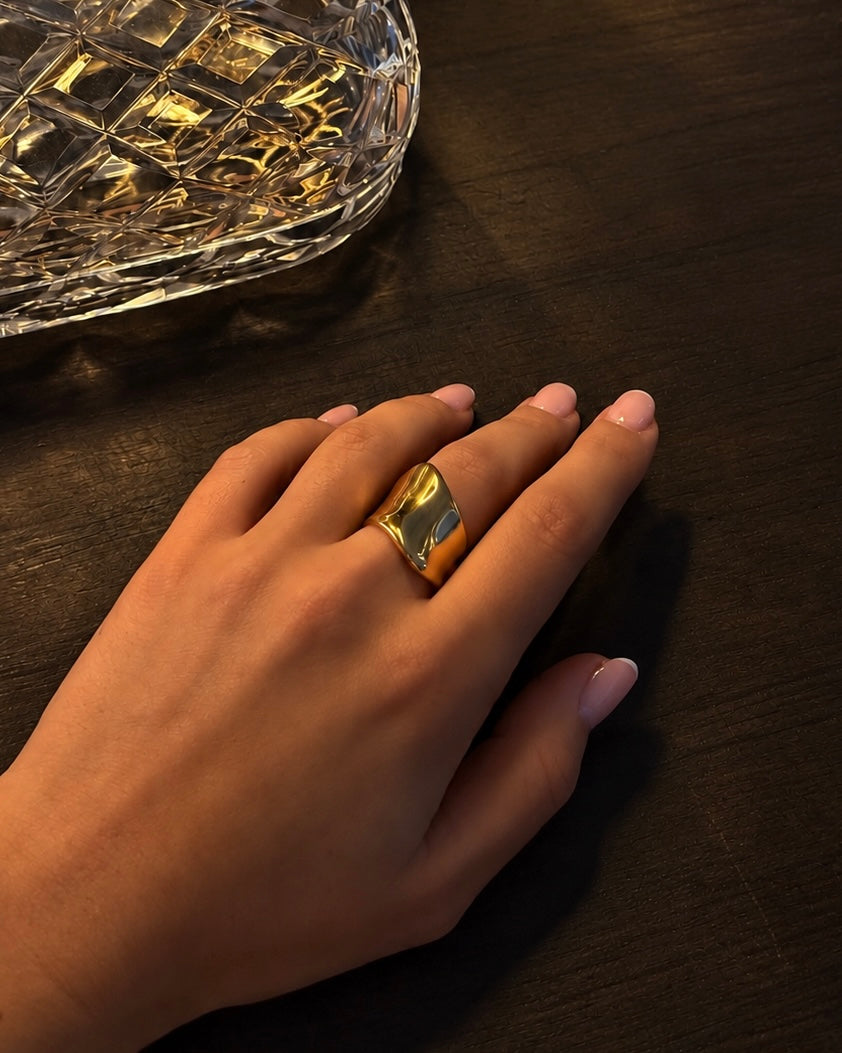Violet Volume Ring i guld buren på modell bredvid en kristallskål – skulpterad statement-ring med vågig, högblank yta. Vattentålig ring i rostfritt stål med 18K guldplätering från Alora Avenue.