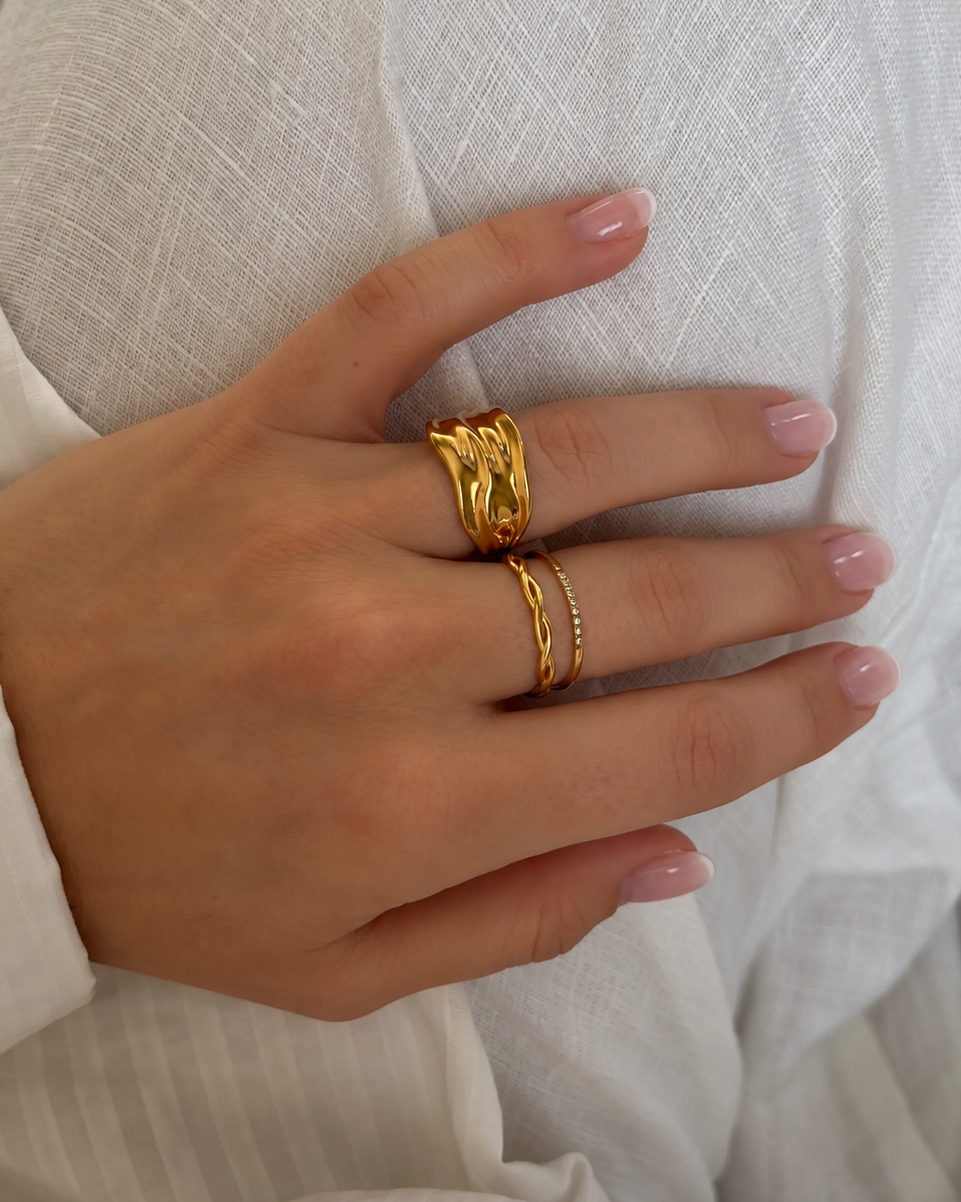Wavy Winter Ring i guld på modell mot vit linneväv, buren tillsammans med en smal stenring. Vattentålig och allergivänlig ring i rostfritt stål med 18K PVD-guldplätering från Alora Avenue.