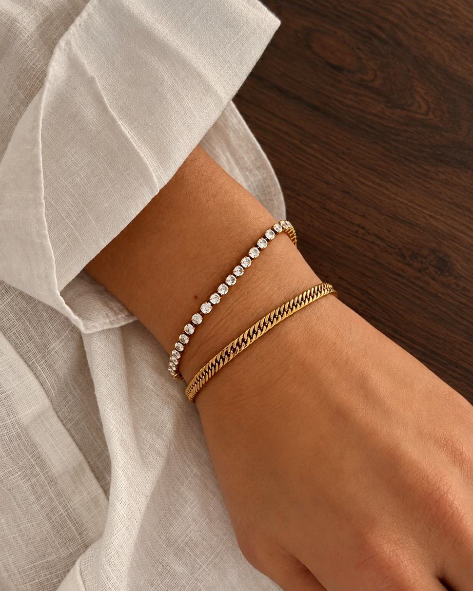 Zion Snow Bracelet i guld på modell, buret tillsammans med ett platt länkarmband mot ett mörkt träbord. Vattentåligt och allergivänligt armband i rostfritt stål med 18K PVD-guldplätering från Alora Avenue.