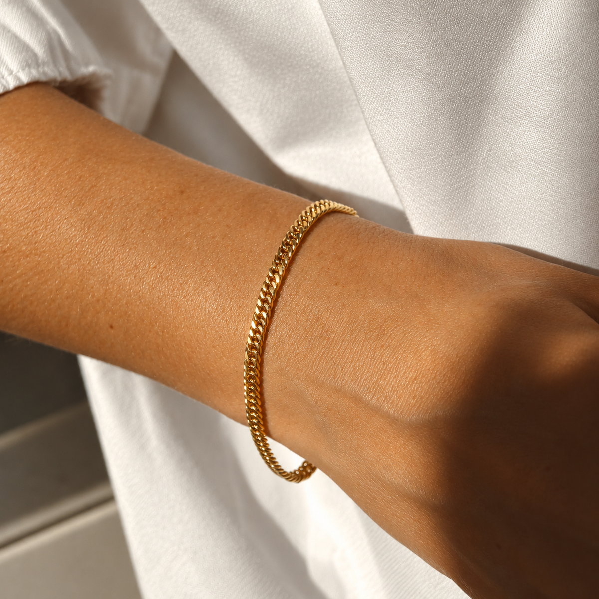 Zion Snow Bracelet i guld buret på modellens handled i varmt solljus. Vattentåligt och allergivänligt armband i rostfritt stål med 18K PVD-guldplätering från Alora Avenue.
